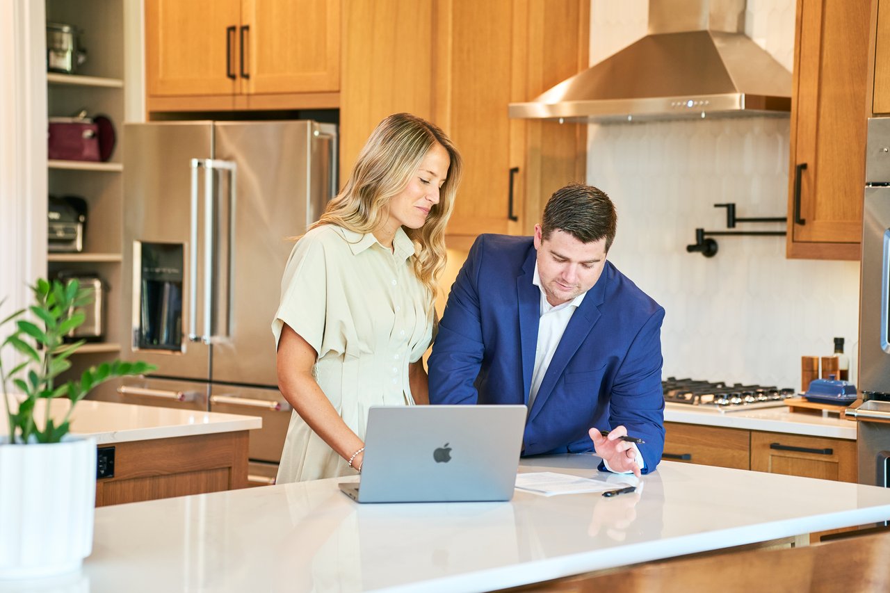 Two real estate agents review paperwork beside a laptop at a kitchen island.