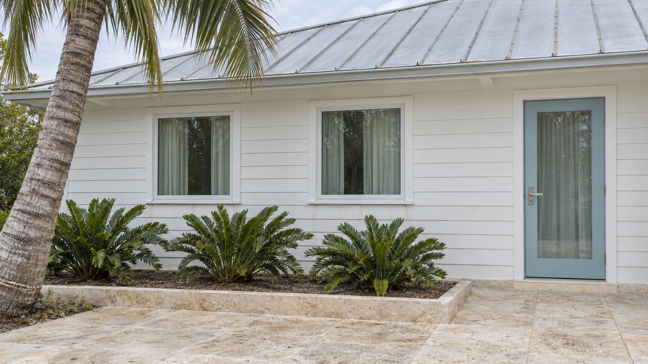 White-sided coastal home with silver metal roof, sea-glass blue door and limestone garden terrace beneath a coconut palm.