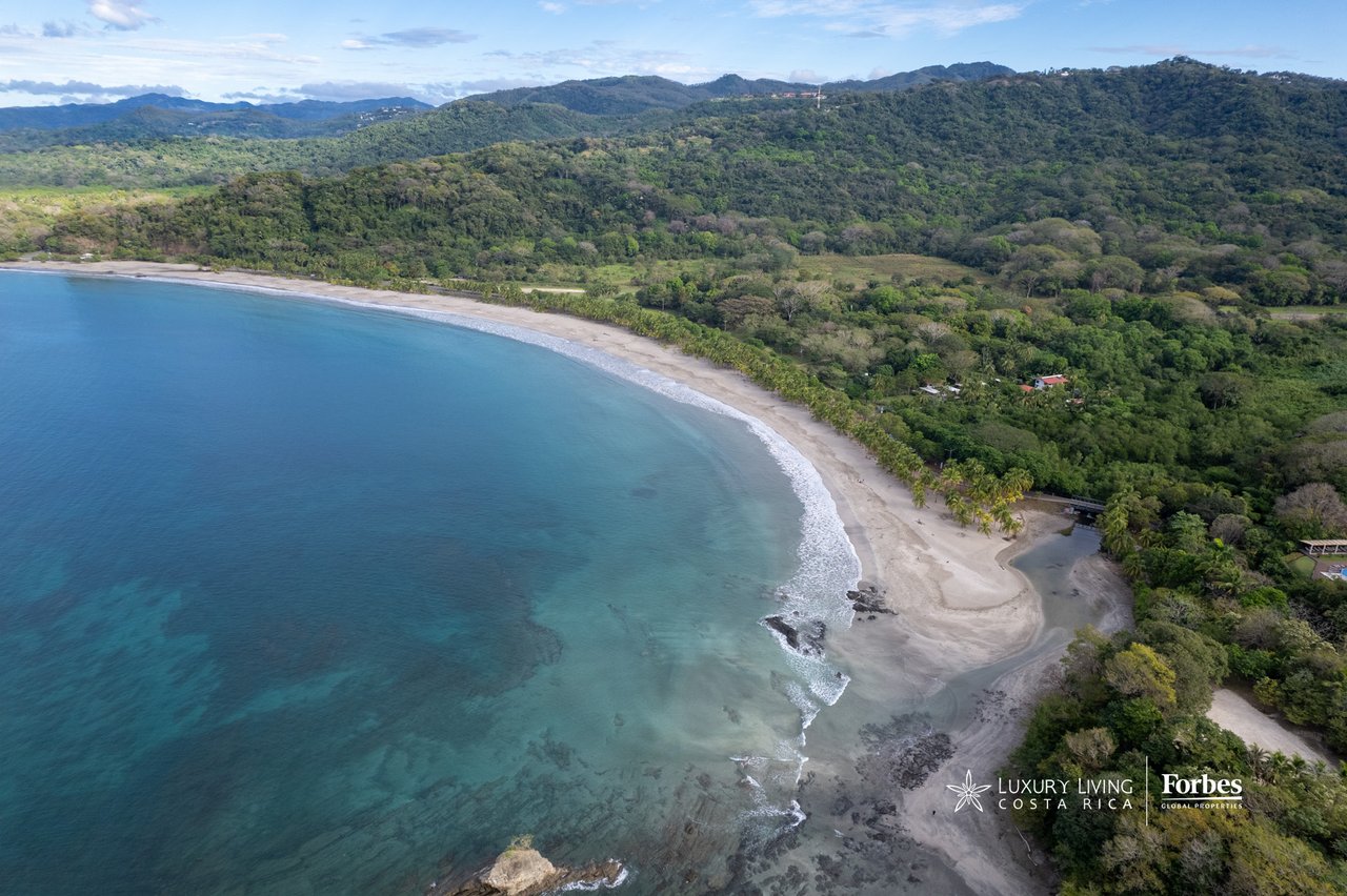 Aerial view of a curved tropical beach, turquoise water, and forested hills.