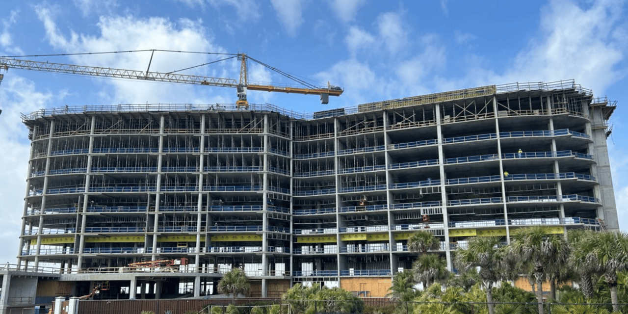 Multi-story concrete building under construction with a crane and palm trees in front.