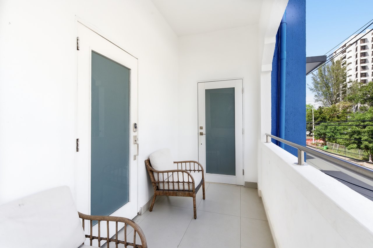Apartment balcony with wicker chairs, frosted-glass doors, and a view of trees and a neighboring high-rise.