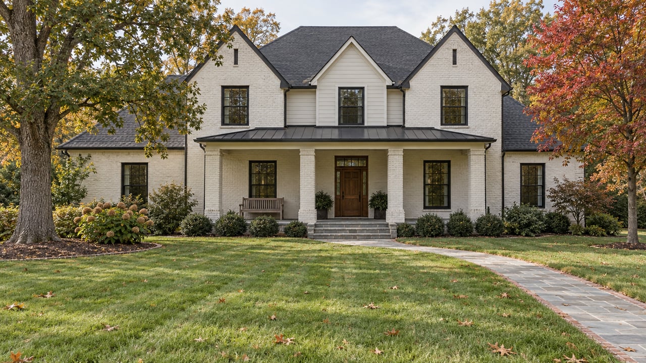 Light-painted brick home with a broad covered porch beyond a deep lawn, framed by mature oak and red maple trees in early fall.