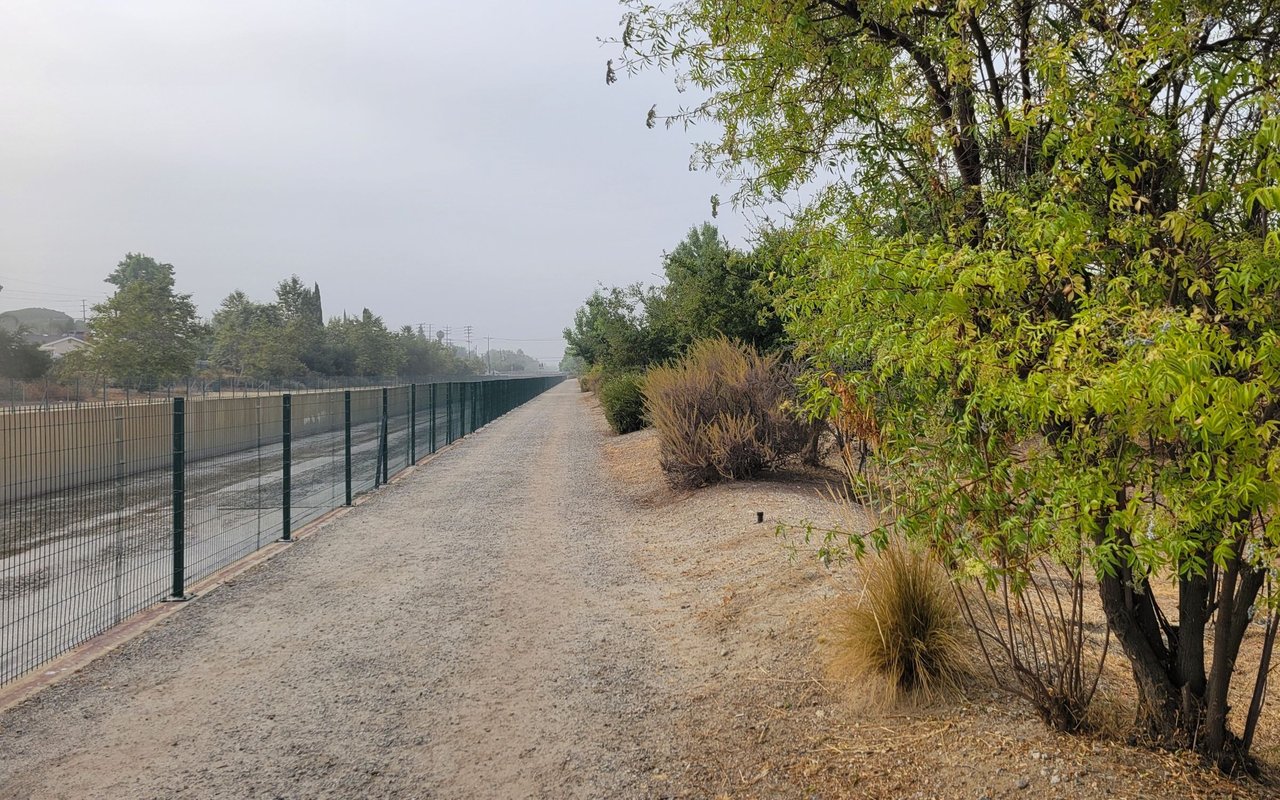 Gravel trail beside a fenced drainage channel, lined with trees and shrubs.