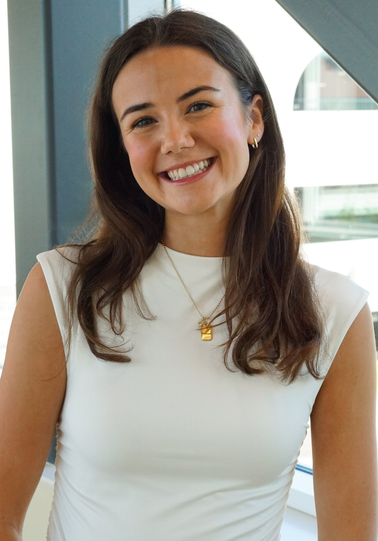 Julia Gray smiling in a white sleeveless top, with long dark hair and gold jewelry.