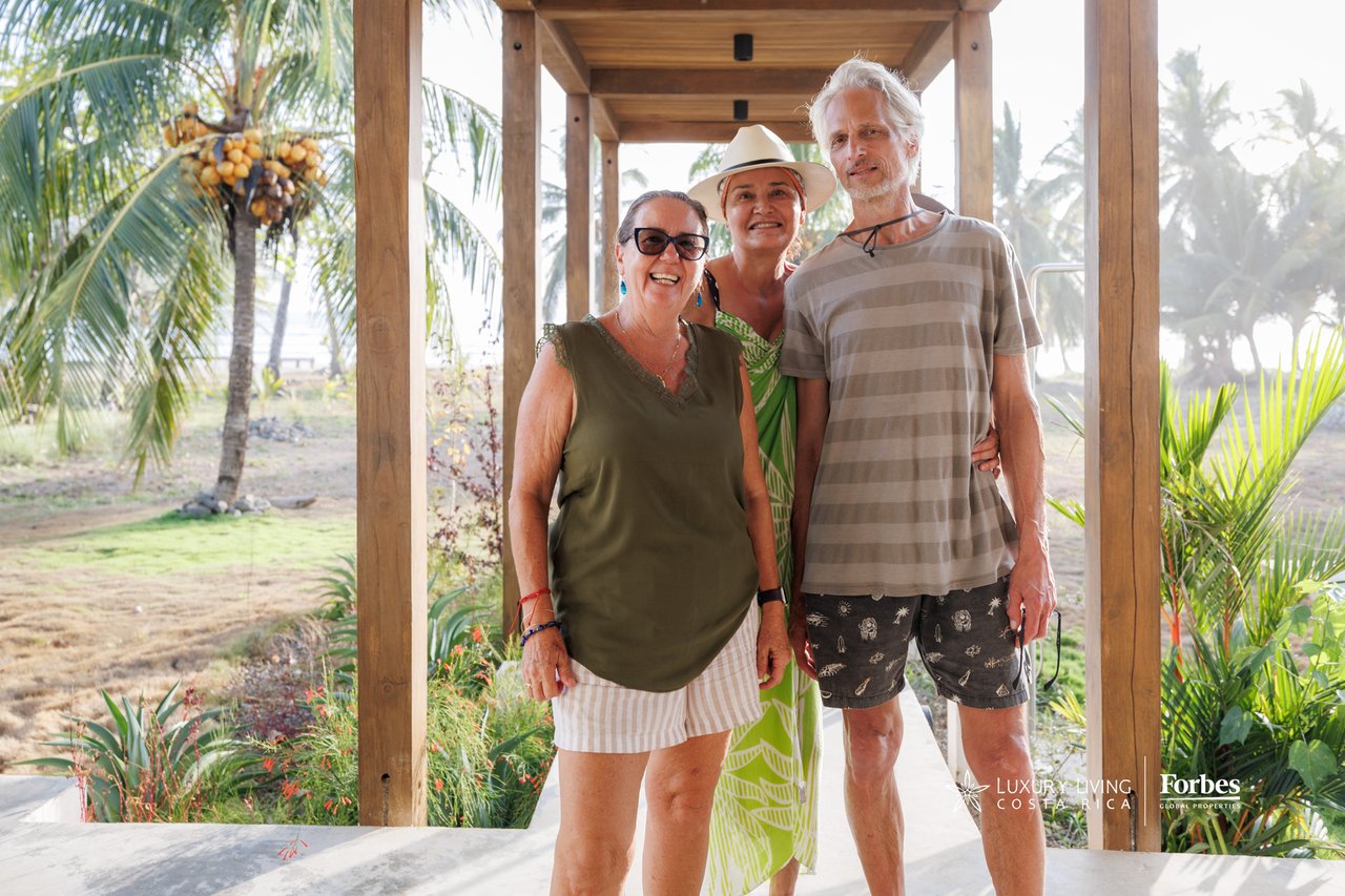 Three adults pose beneath a wooden pergola surrounded by tropical landscaping.