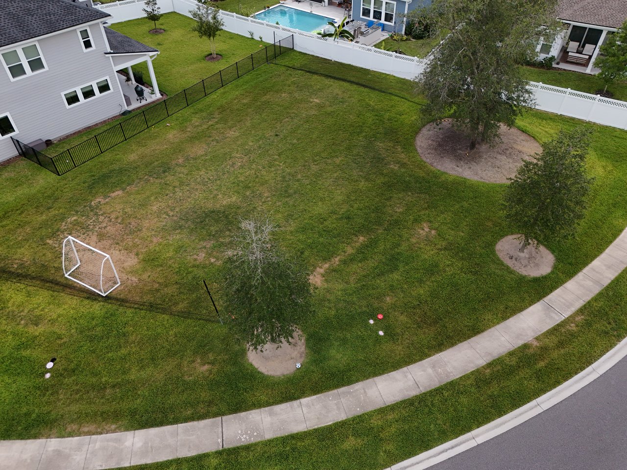 Aerial view of a broad grassy yard with scattered trees, a soccer goal, and a black fence.
