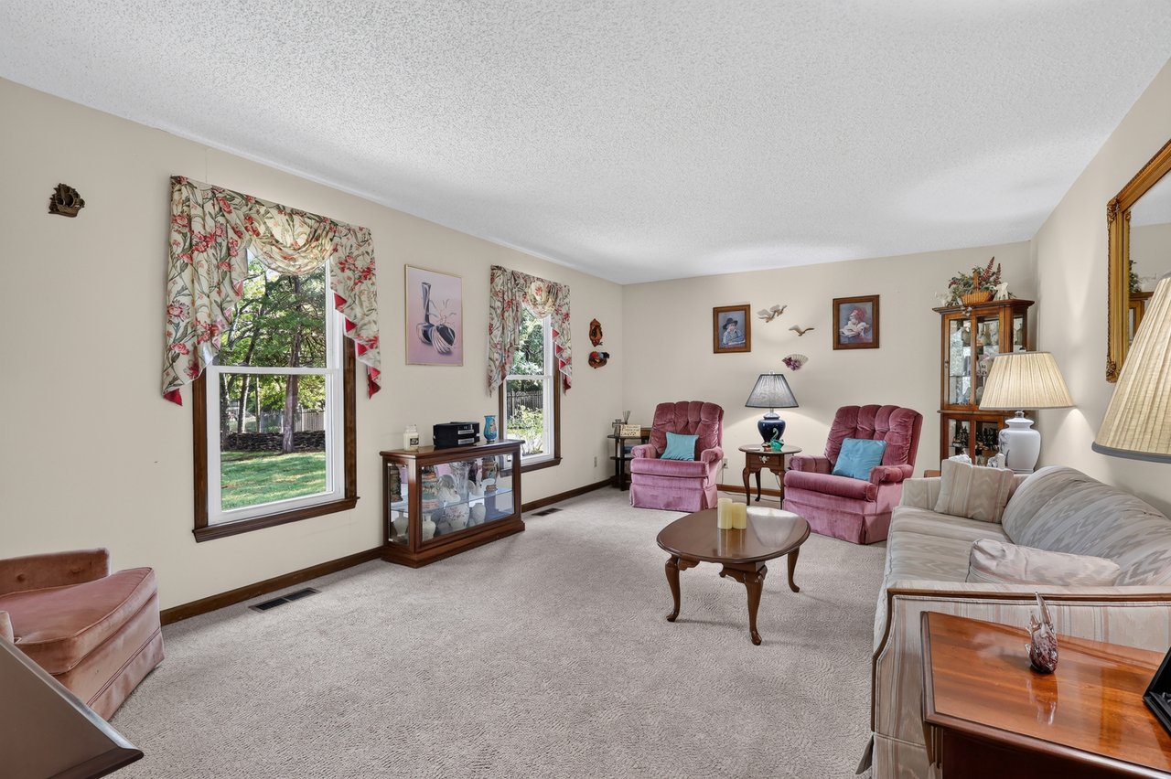 Furnished living room with pink recliners, patterned curtains, and large windows overlooking a leafy yard.