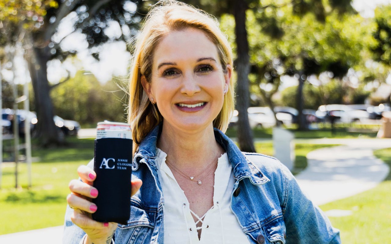 A smiling woman holds up a black koozie with a logo in a park, wearing a white shirt and denim jacket.