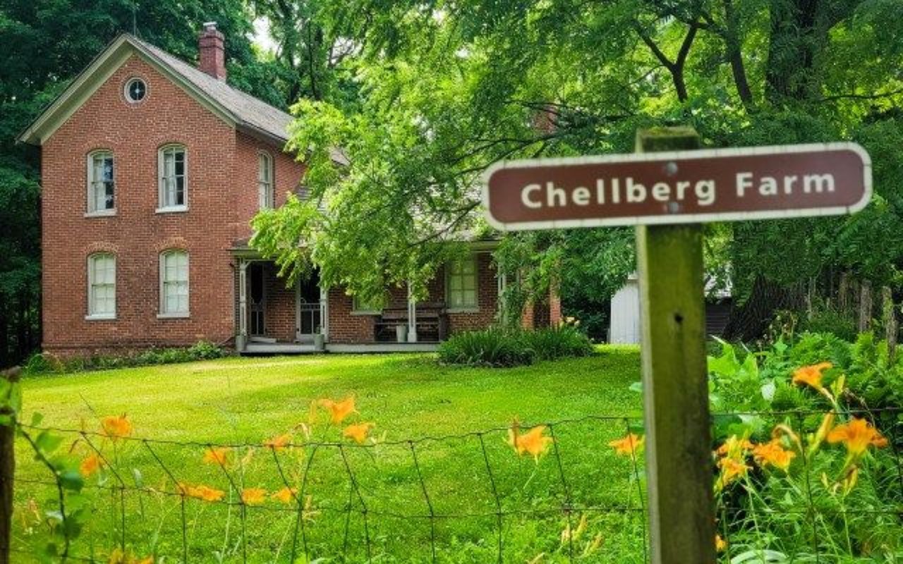 Red-brick farmhouse behind a Chellberg Farm sign, framed by leafy trees and orange flowers.