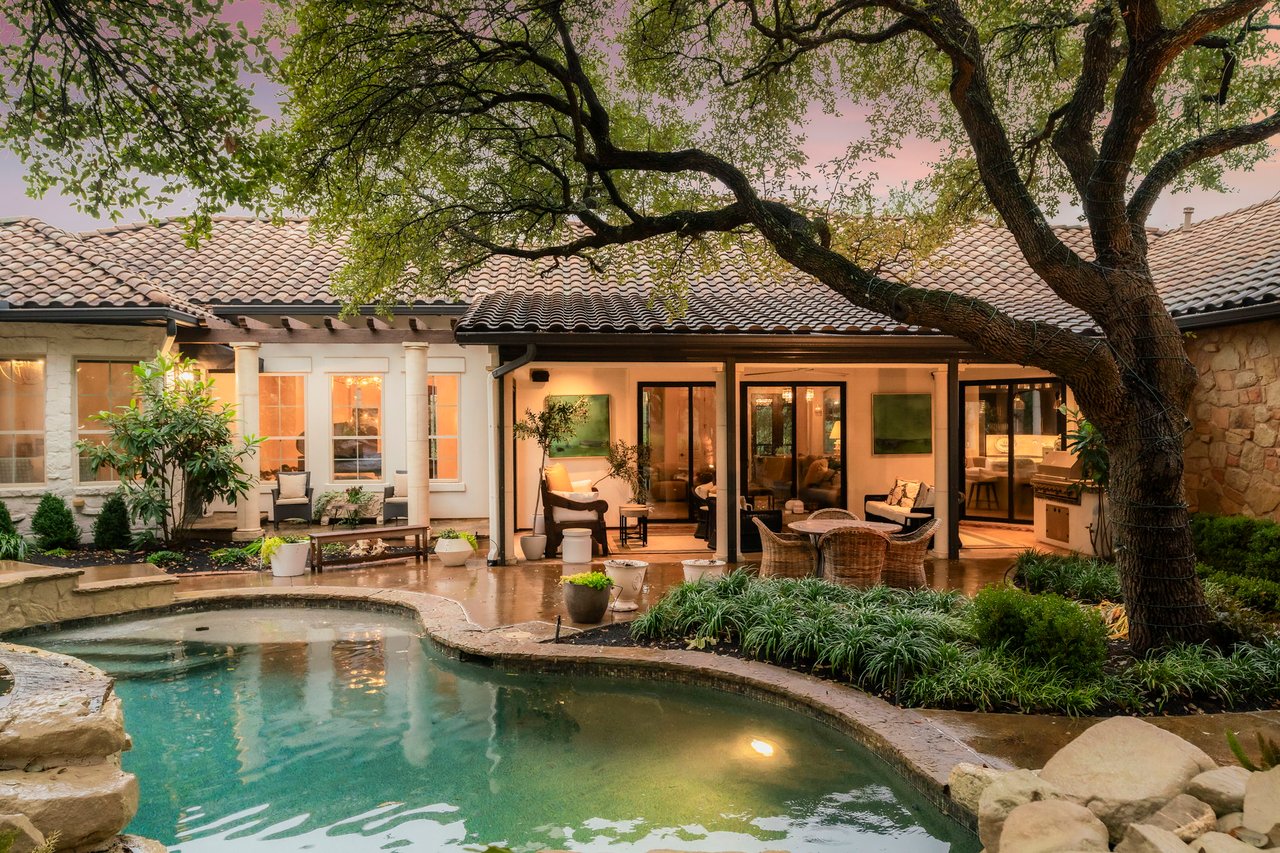 Stone home with a covered patio and pool beneath a sprawling tree at dusk.
