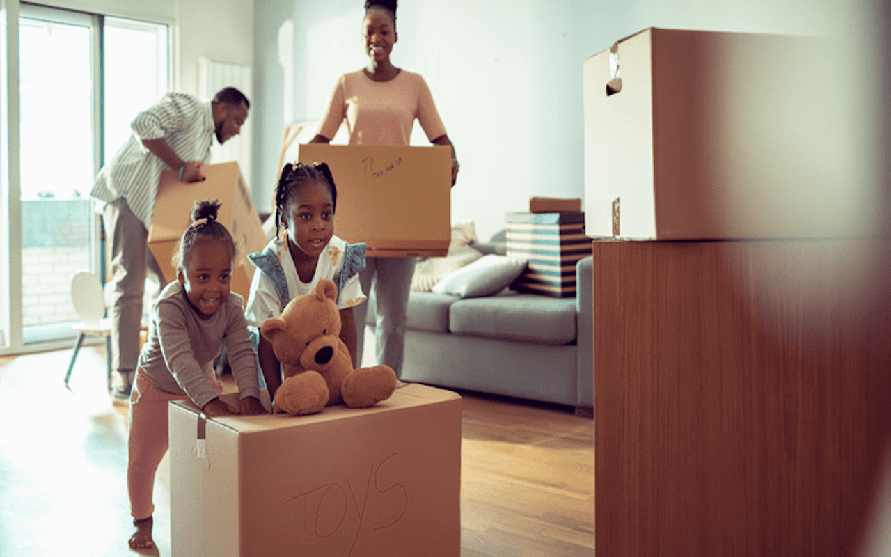 Happy family with two young children pushing cardboard moving boxes in a bright living room.