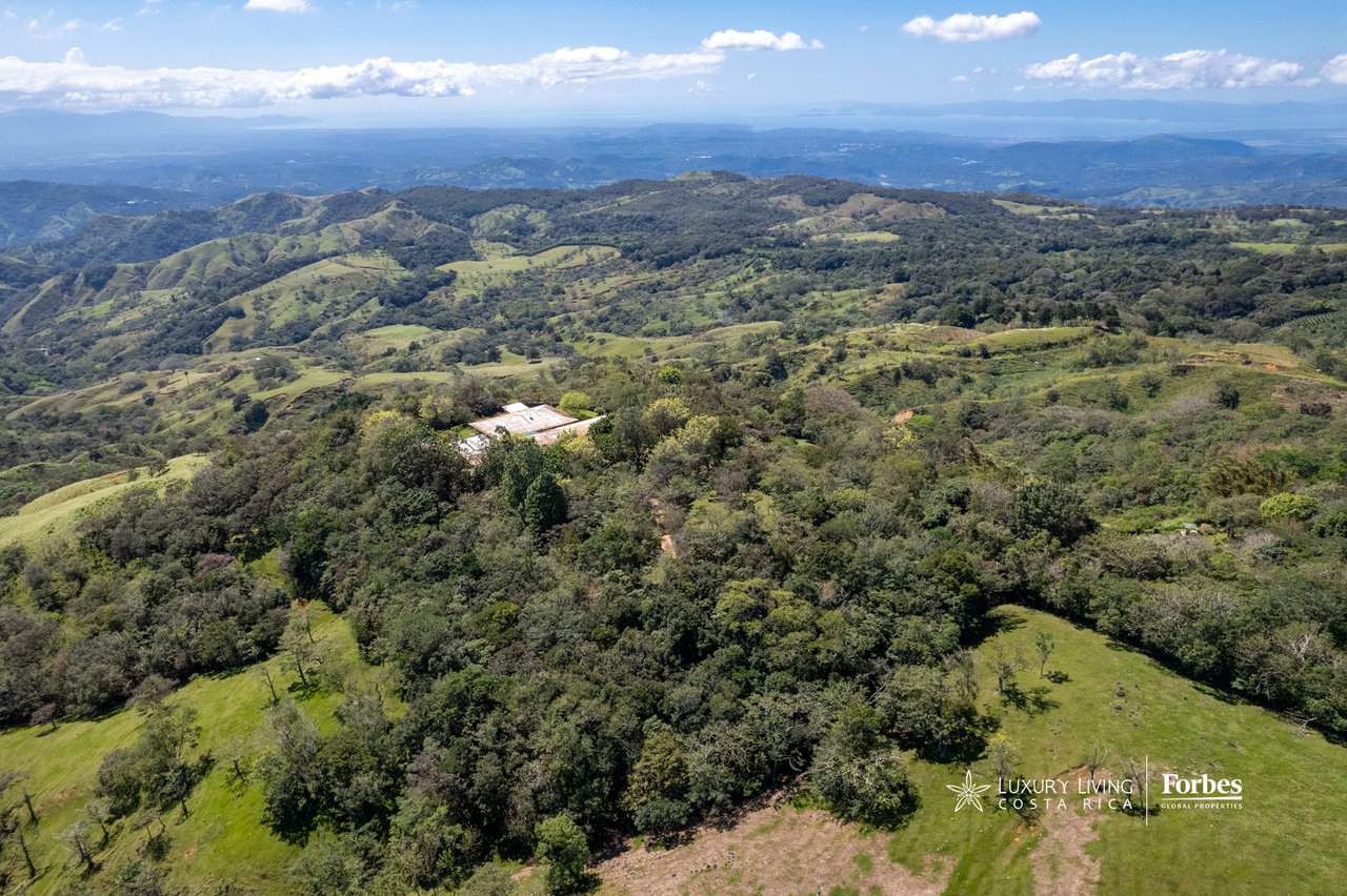 Aerial view of a secluded property amid forested hills, with expansive mountain ranges on the horizon.