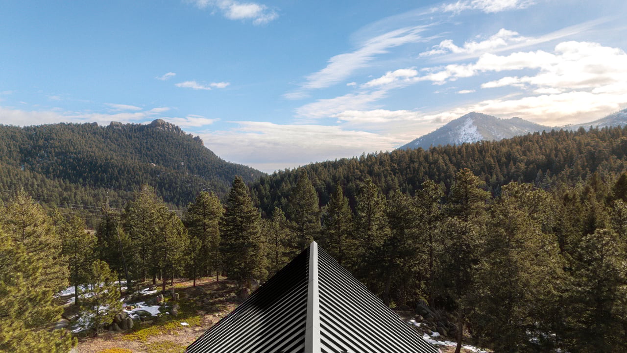 Views of Boulder's Flatirons and snow-capped peaks over pine forest, with modern metal roof in foreground