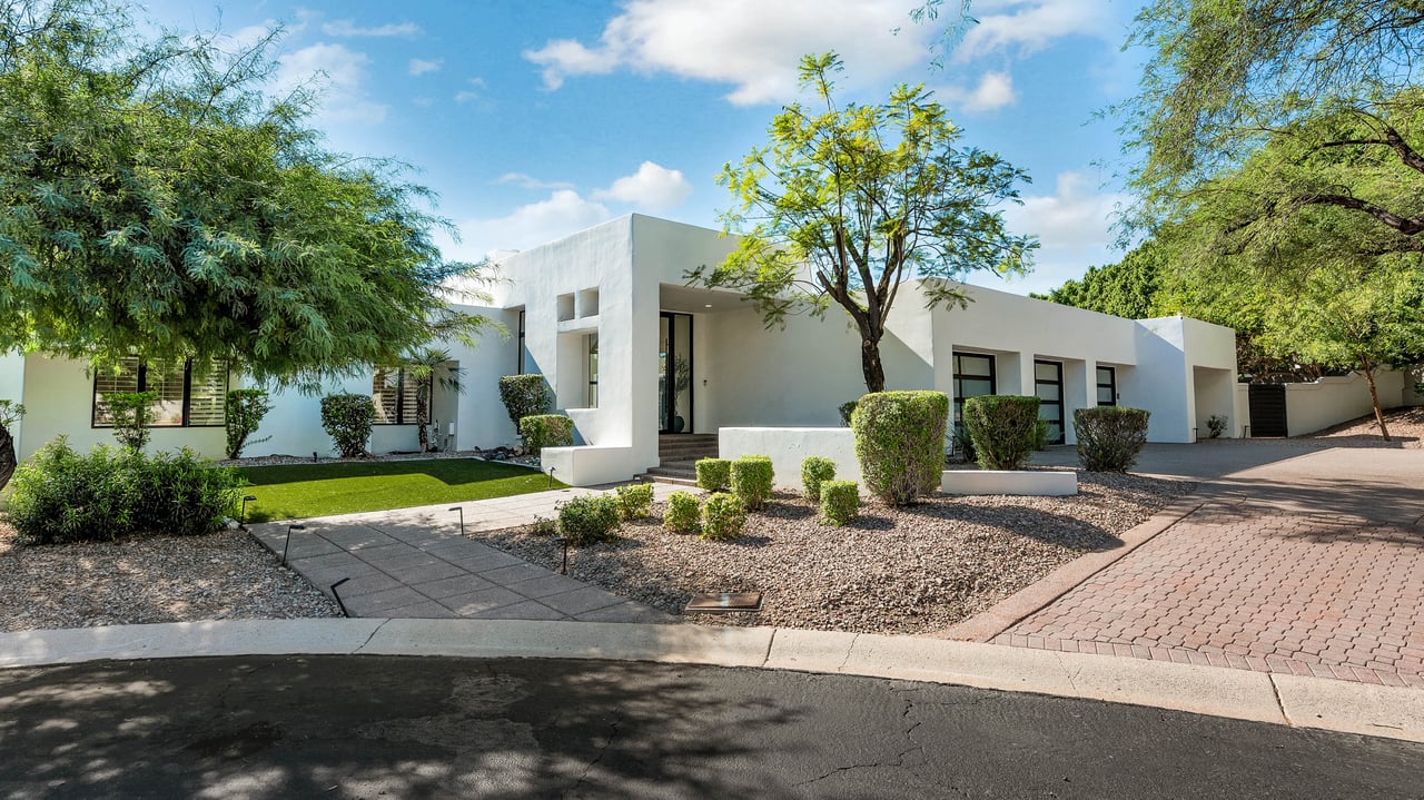 Modern white single-story home with a flat roof, mature trees, gravel landscaping, and a paved driveway.