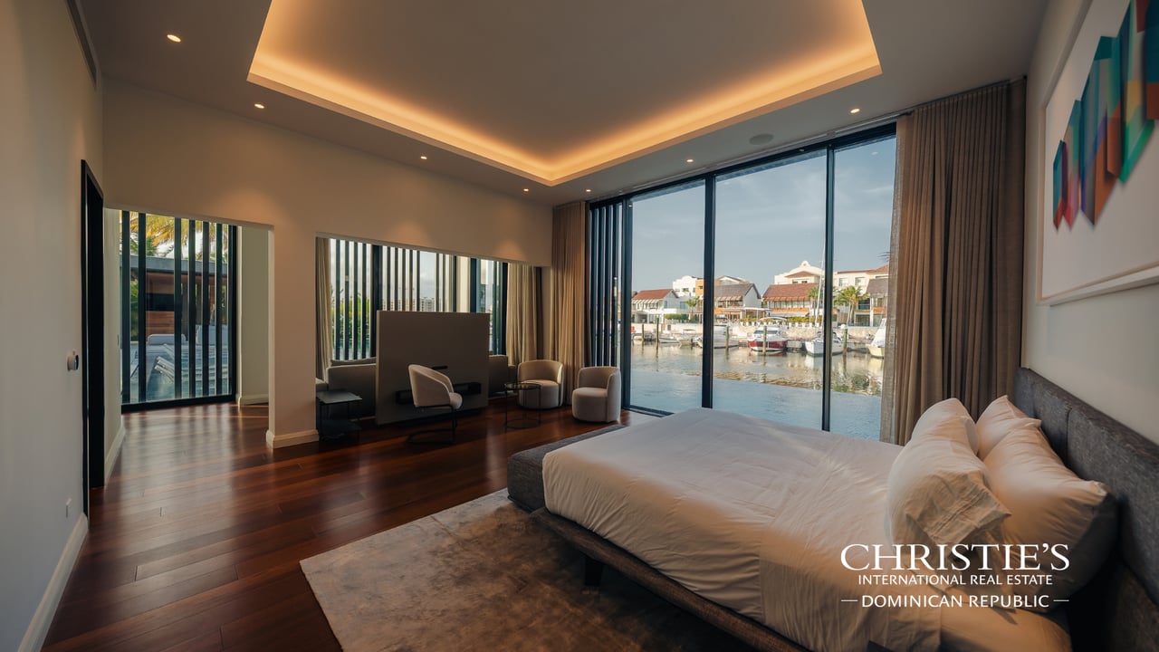 Furnished bedroom with floor-to-ceiling windows overlooking marina water and waterfront homes.