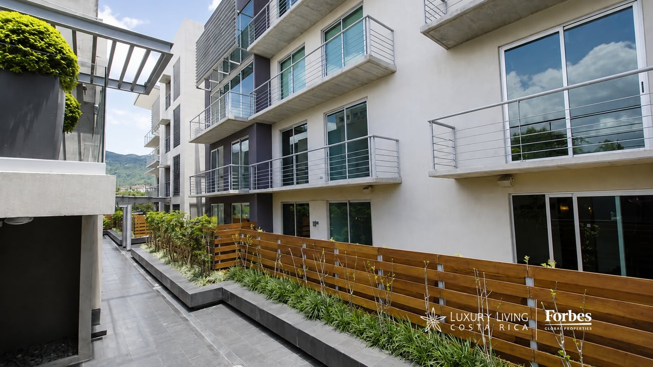 Modern apartment building courtyard with balconies, raised planters, and distant green mountains.