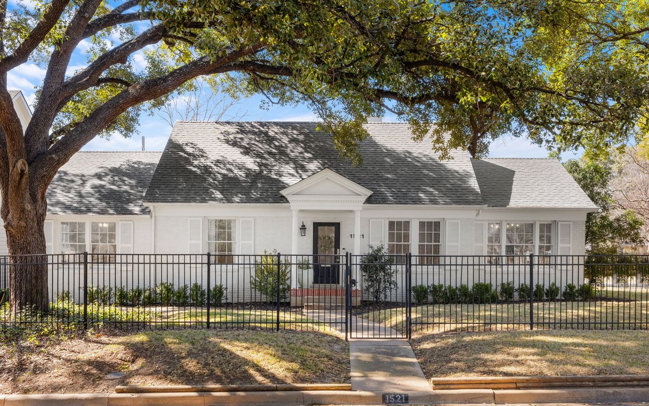 White single-story home behind a black metal fence, shaded by a large oak tree.