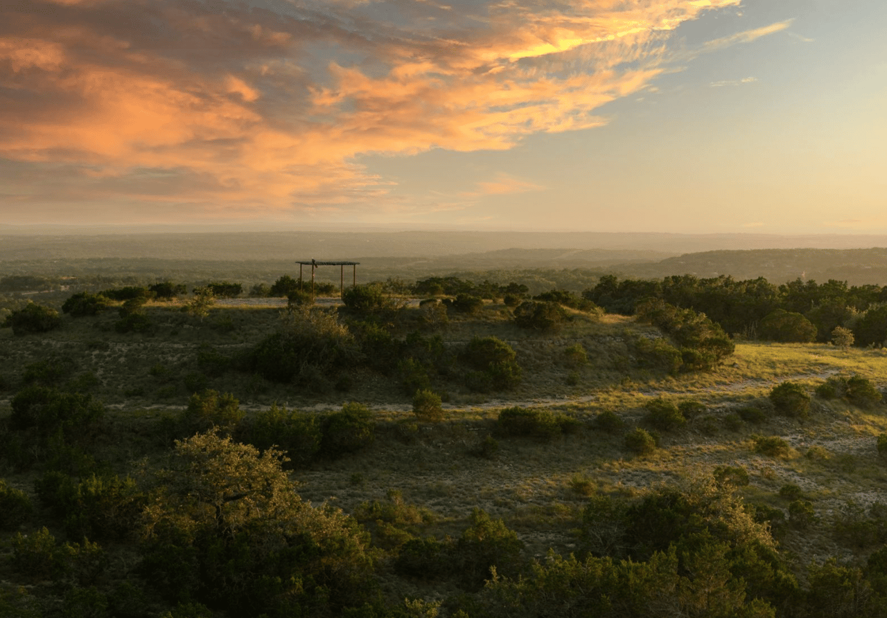 Rugged ranchland and distant wooded hills beneath orange sunset clouds, with a wooden frame on the ridge.