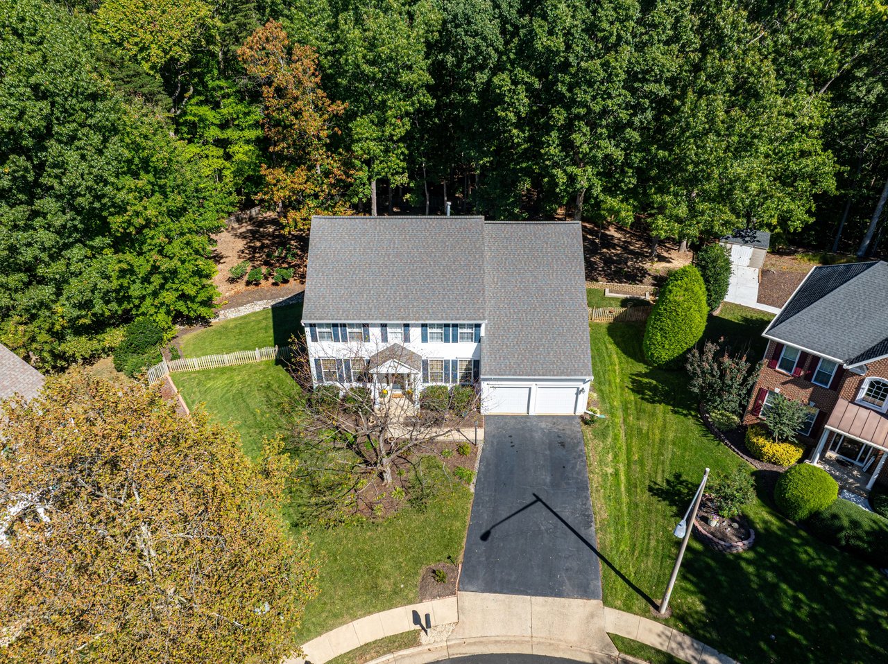 Aerial view of a two-story home with a long driveway, lawn, and wooded backyard.