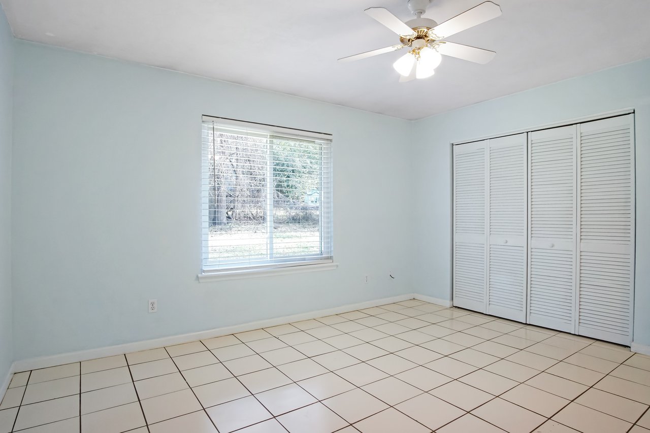 Empty room with pale blue walls, tiled floor, large window with blinds, and a ceiling fan. White closet doors, bright, minimalistic feel.