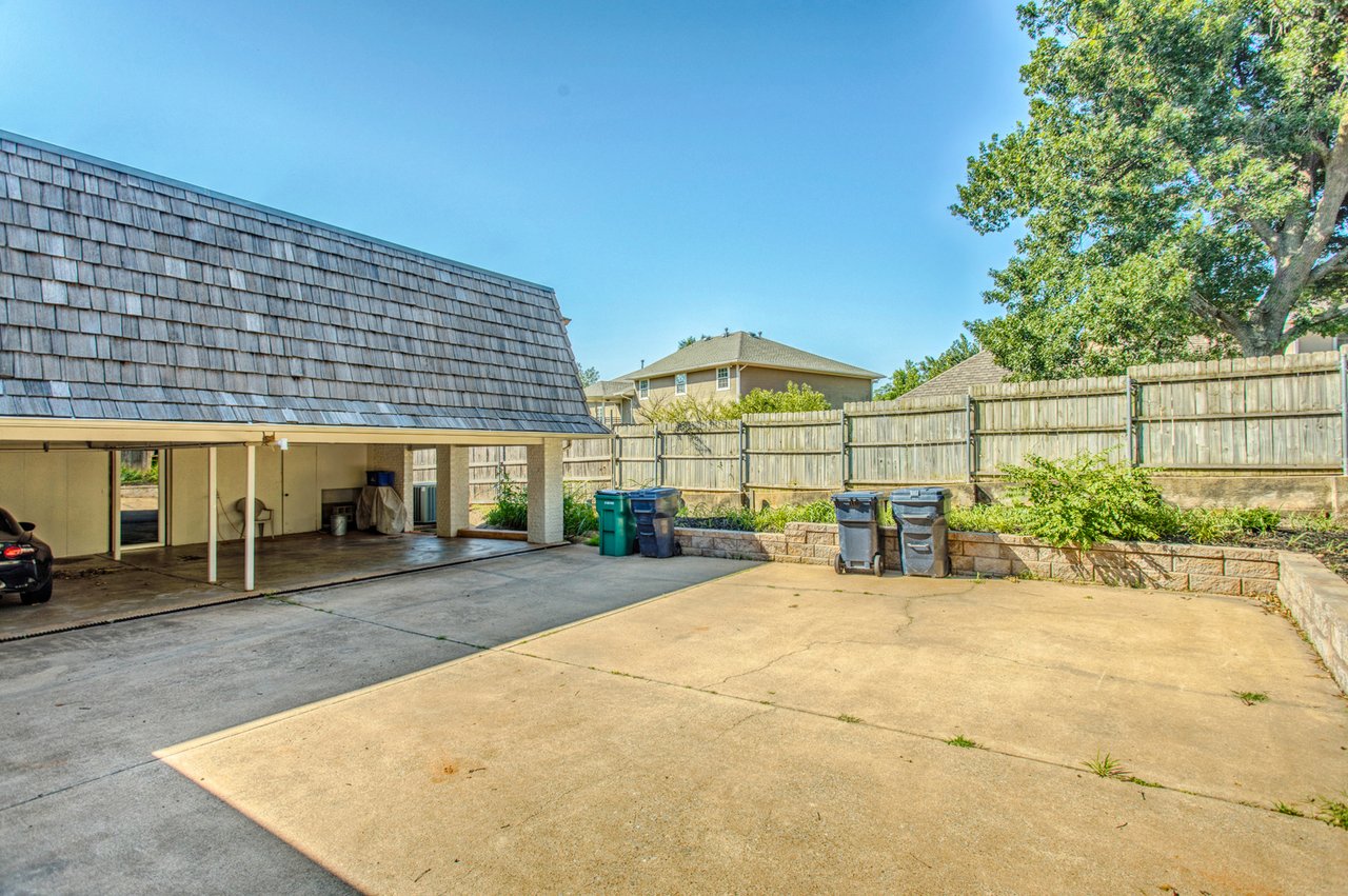 Concrete driveway beside a covered carport, with a wooden privacy fence and raised planting bed.