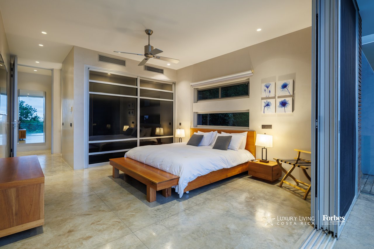 Modern bedroom with a low wooden bed, dark sliding closet doors, and large windows.