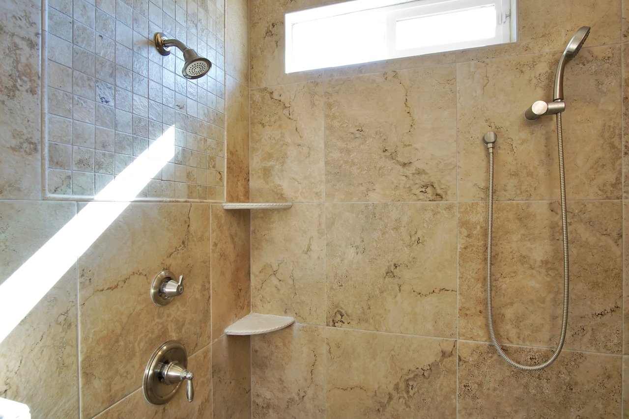 Spacious beige-tiled shower with two showerheads, one fixed and one handheld, under bright natural light from a narrow window above.