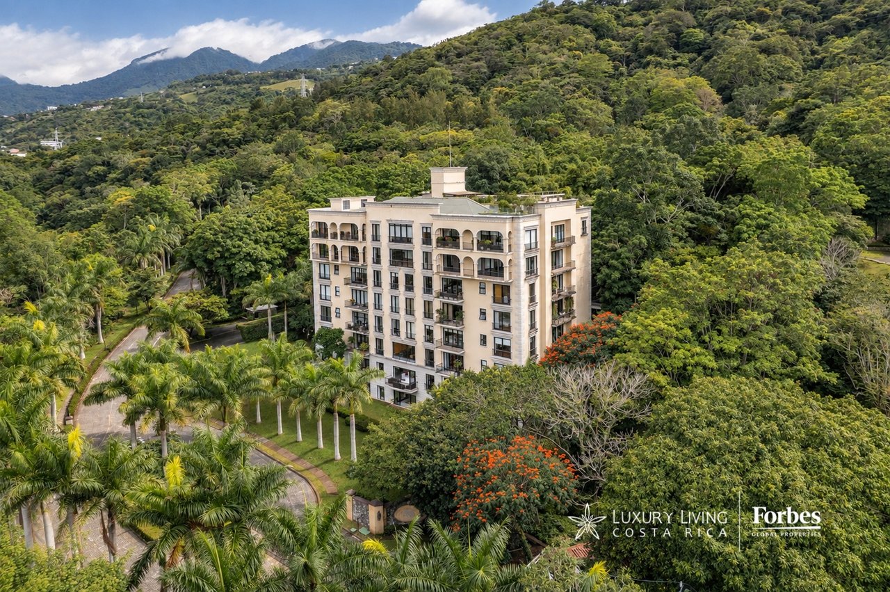 Aerial view of a cream-colored apartment building amid tropical greenery, with forested mountains behind it.