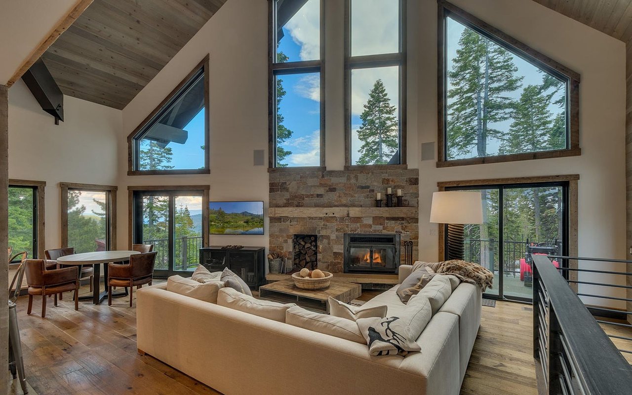 Living room with vaulted wood ceiling, stone fireplace, and tall windows overlooking trees.