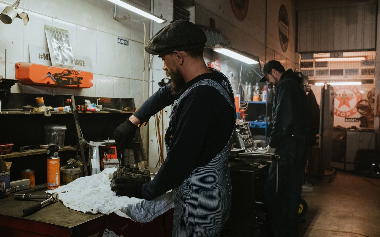 Mechanic in striped overalls works on a part at a cluttered workshop bench.