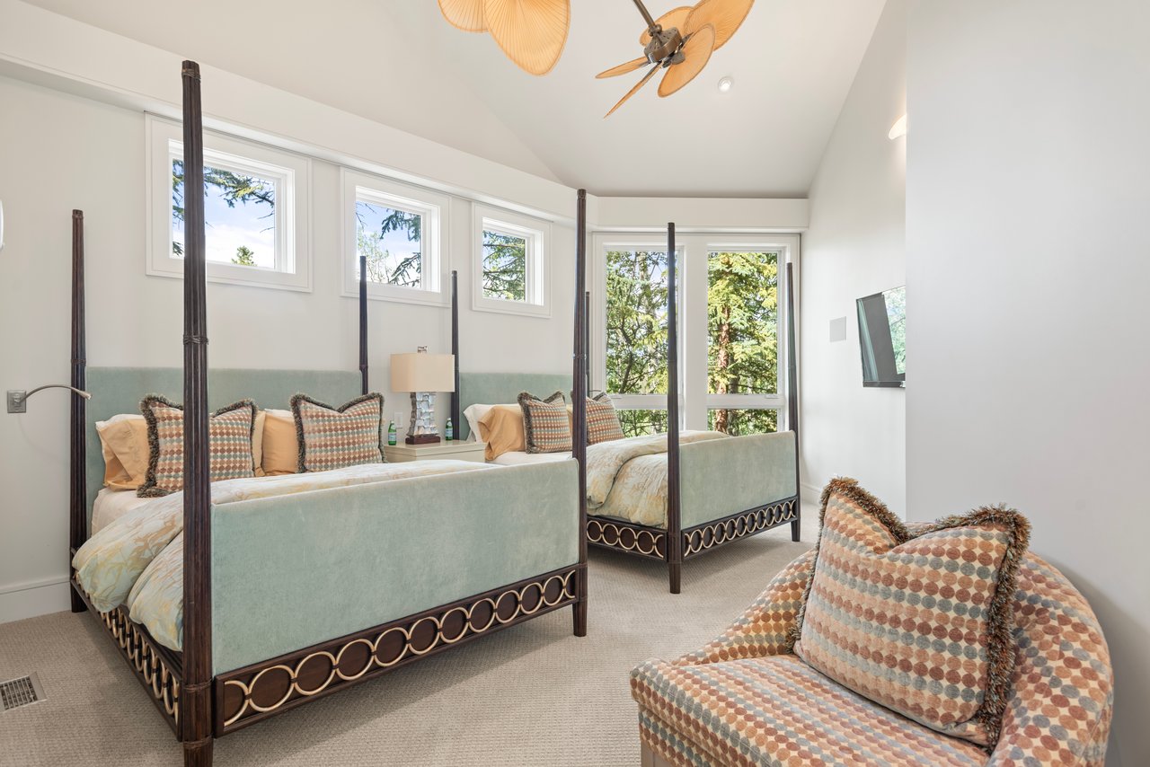 Bedroom with two pale green four-poster beds and tall windows overlooking trees.