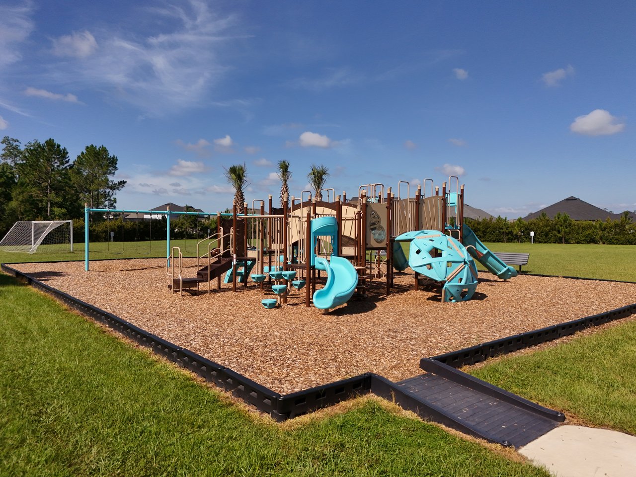 Community playground with turquoise slides and climbing equipment beside a grassy field and soccer goal.