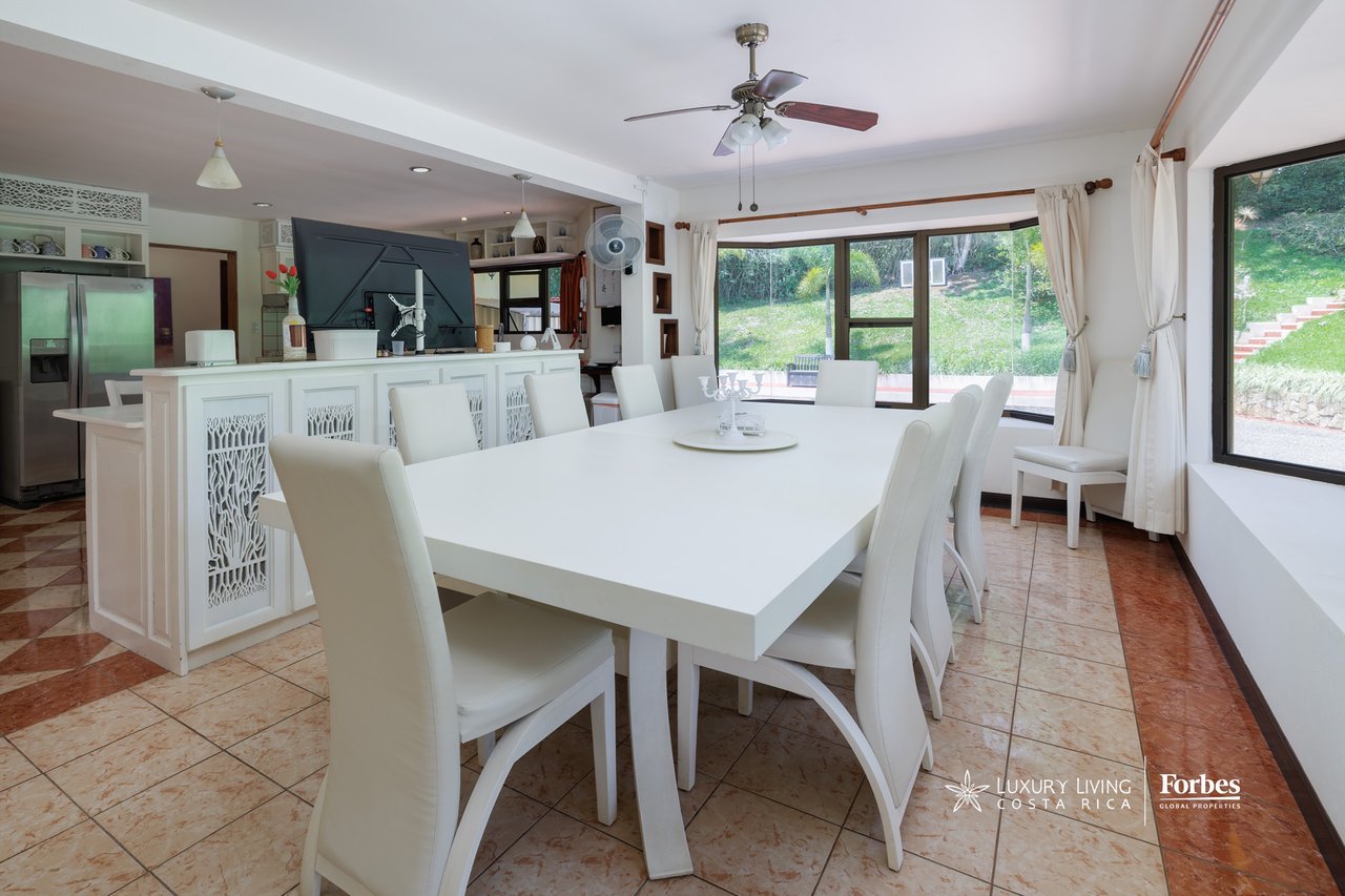 Dining room with a large white table beside a kitchen bar and garden-facing windows.