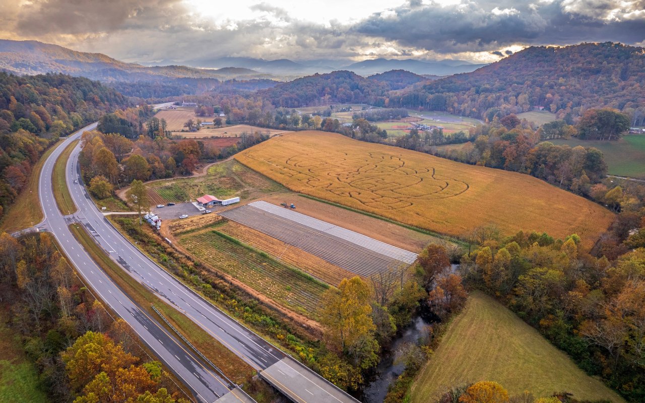 Autumn farm with a large corn maze, fields, and wooded mountains beside a curving highway.