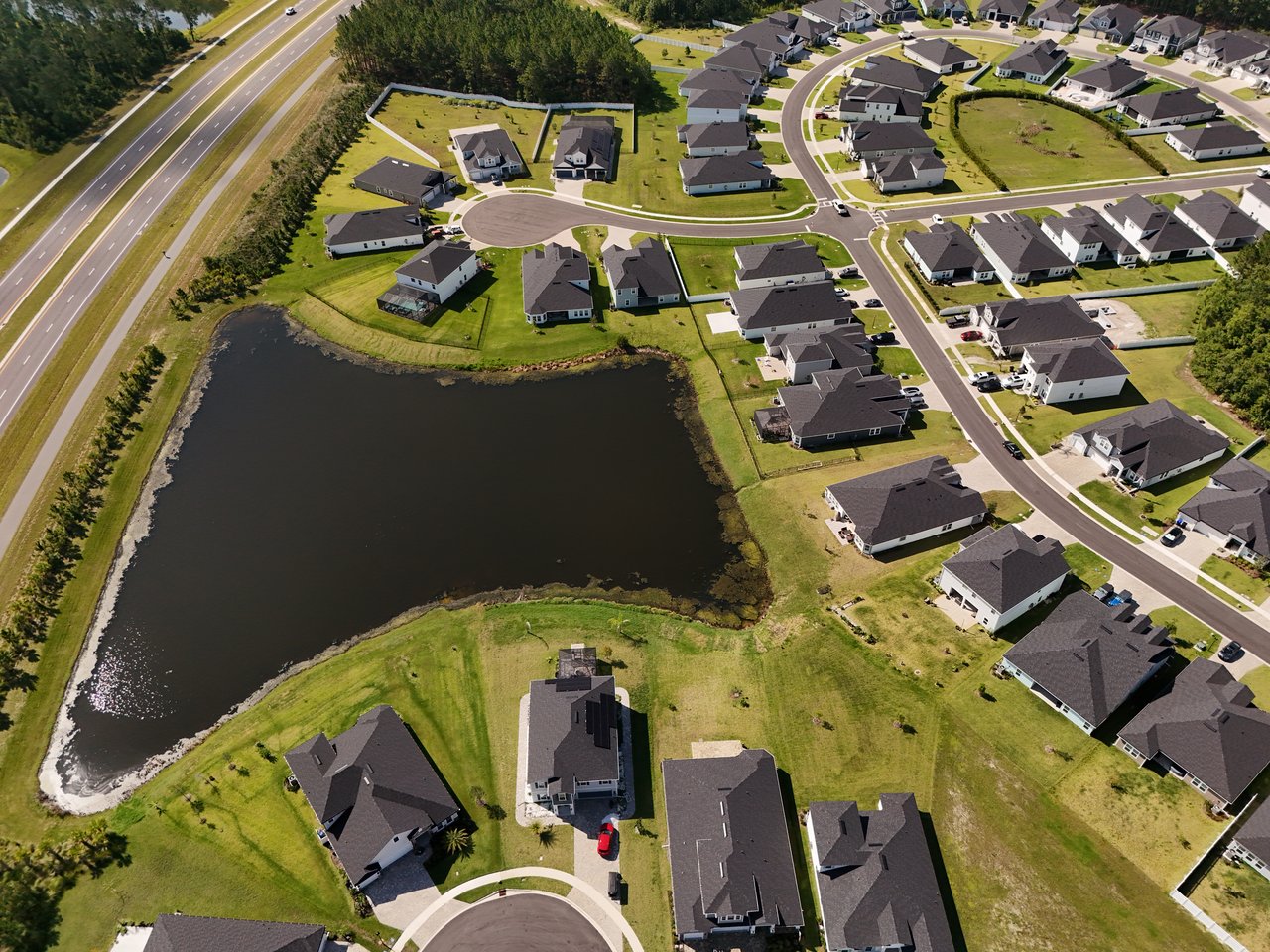 Aerial view of homes around a large pond, with a highway bordering the neighborhood.