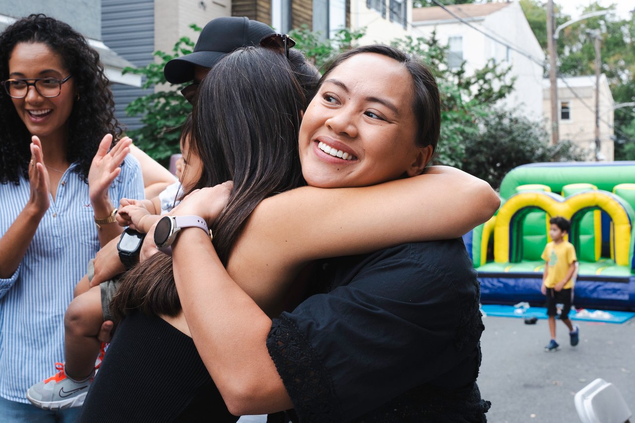 Neighbors embrace at an outdoor block party beside a colorful bounce house.