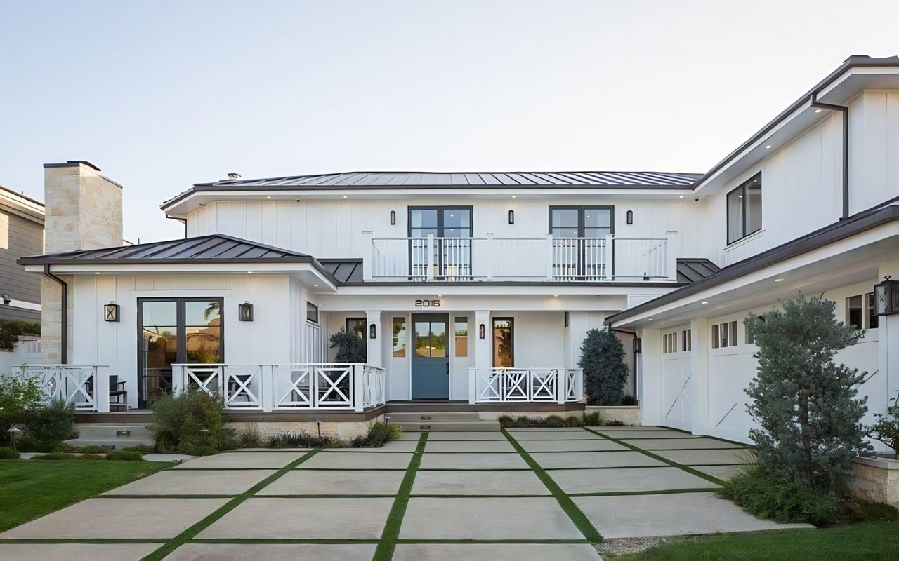  A large, modern white house with a two-car garage, a blue front door, and a driveway with green grass growing in the cracks.
