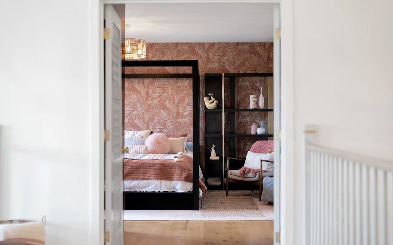 Staged bedroom with a black four-poster bed, blush bedding, and tropical leaf wallpaper.