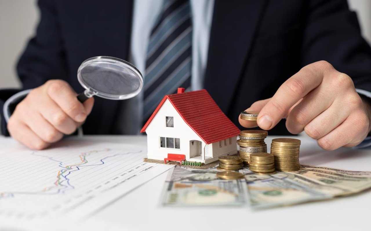 A person adds a coin beside a model house and stacks of coins.