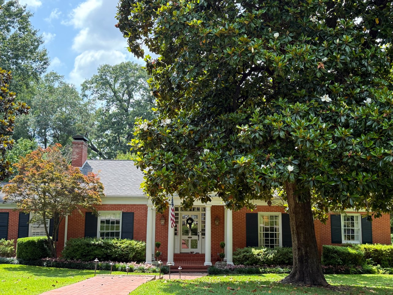 Traditional single-story red brick luxury home in Houston featuring a large southern magnolia tree, black shutters, and a brick walkway, typical of established neighborhoods like River Oaks and Memorial.
