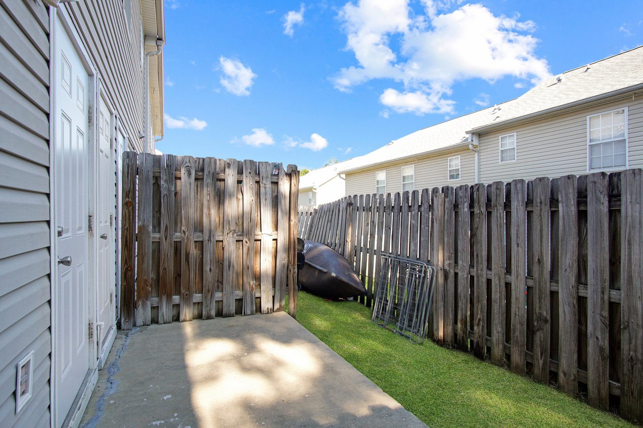 Narrow alley beside beige buildings, enclosed by tall wooden fences. Concrete path with some grass, bright clear sky above, kayak and metal frames stacked nearby.Narrow alley beside beige buildings, enclosed by tall wooden fences. Concrete path with some grass, bright clear sky above, kayak and metal frames stacked nearby.