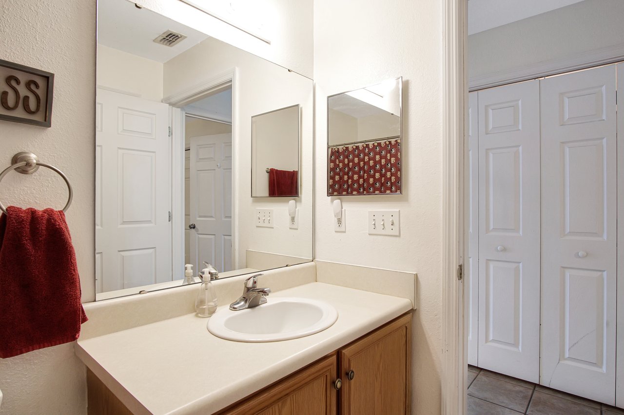 Clean bathroom with wood cabinet, white countertop, and sink. Mirror reflects red towel and patterned curtain. Bright, organized, and functional space.