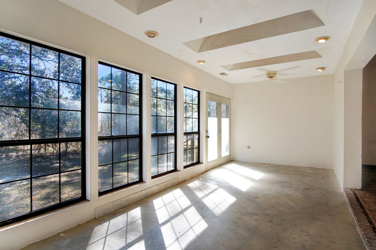 A sunlit room with large windows casting grid-patterned shadows on the concrete floor. It has a white ceiling with recessed lights and a fan.