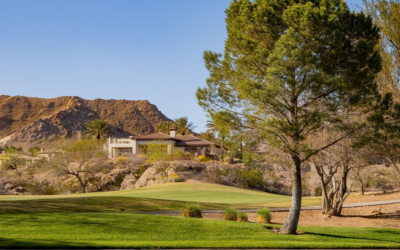 Desert home beside a golf course, backed by rugged mountains and framed by a large pine tree.