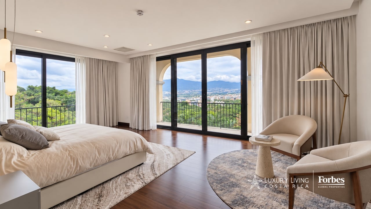 Penthouse bedroom with a spacious bed and sitting area overlooking green hills, the city and distant mountains.