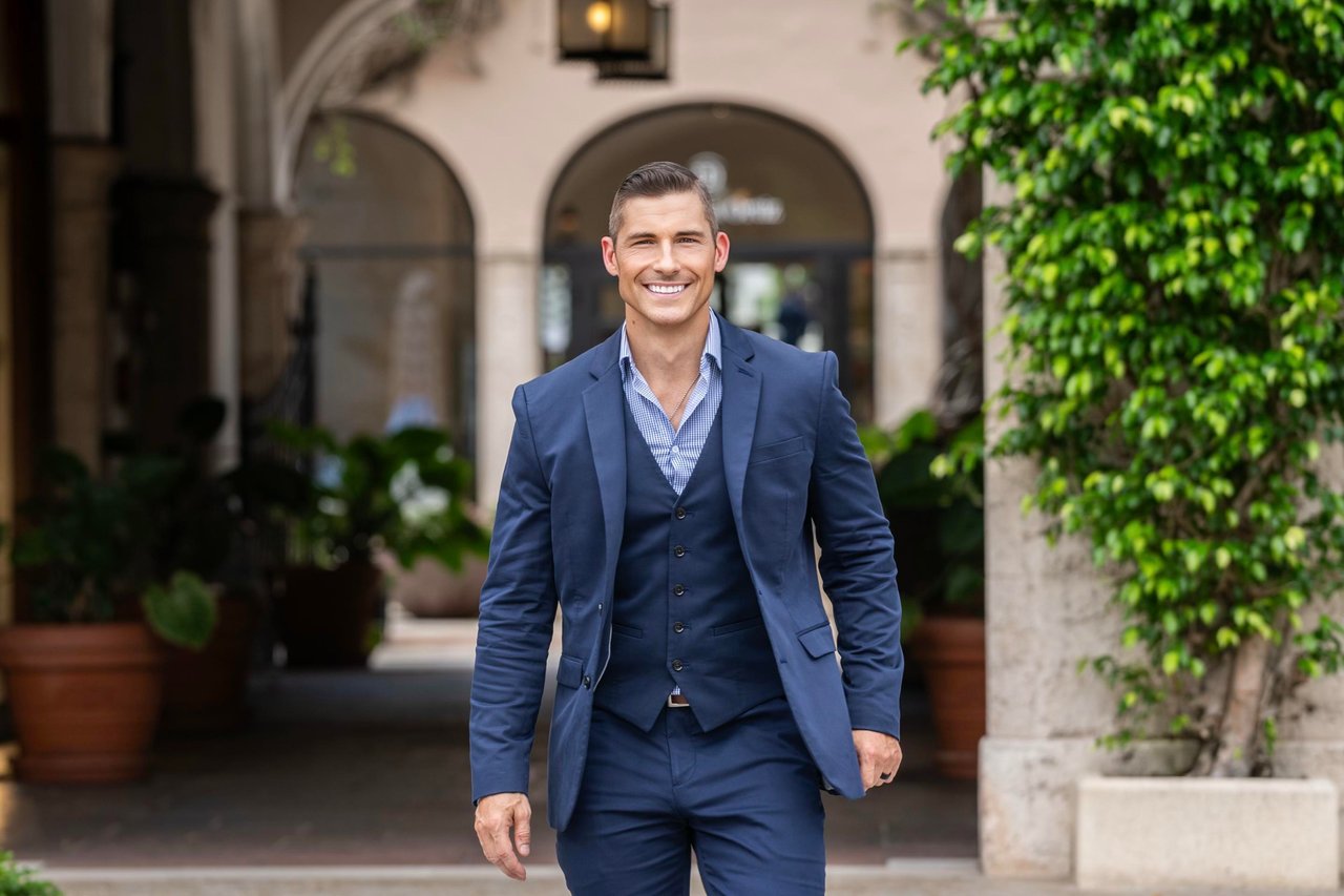 Timothy Mandala in a navy suit, standing in a leafy courtyard with arched entrances.
