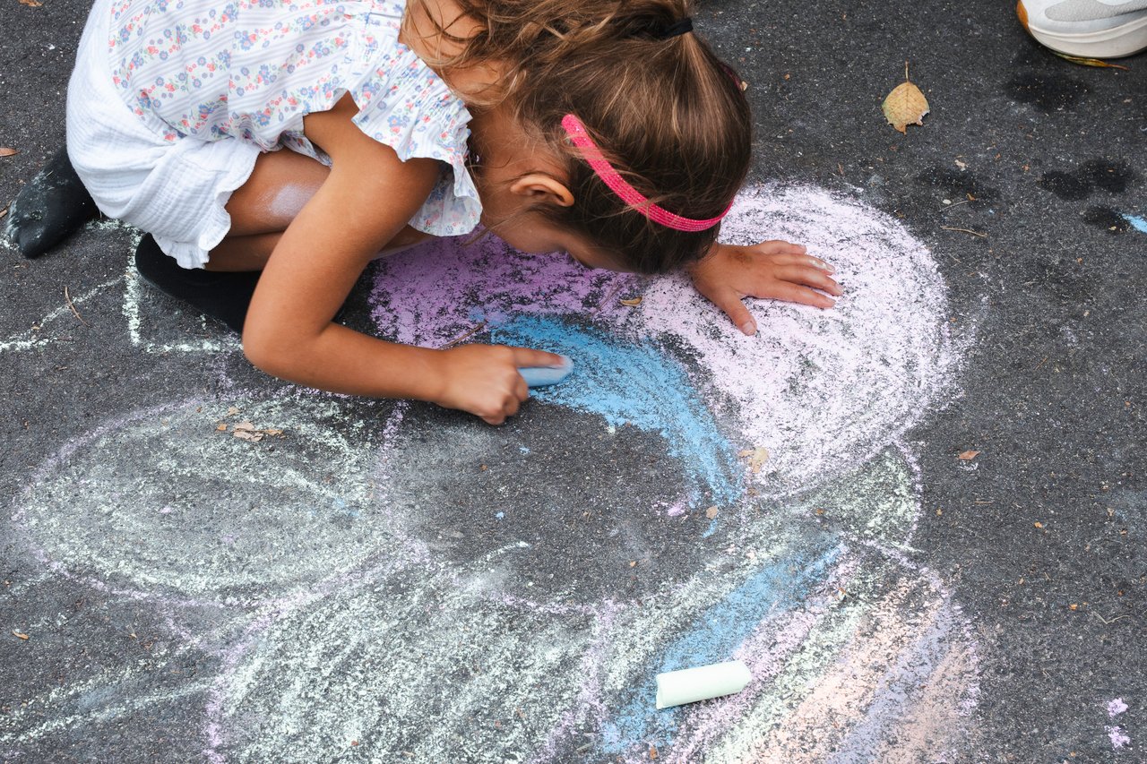A girl crouches over a large, colorful chalk drawing on pavement.