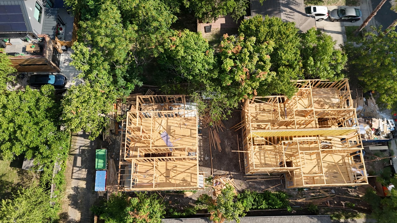 Aerial view of two wood-framed homes under construction among mature trees.