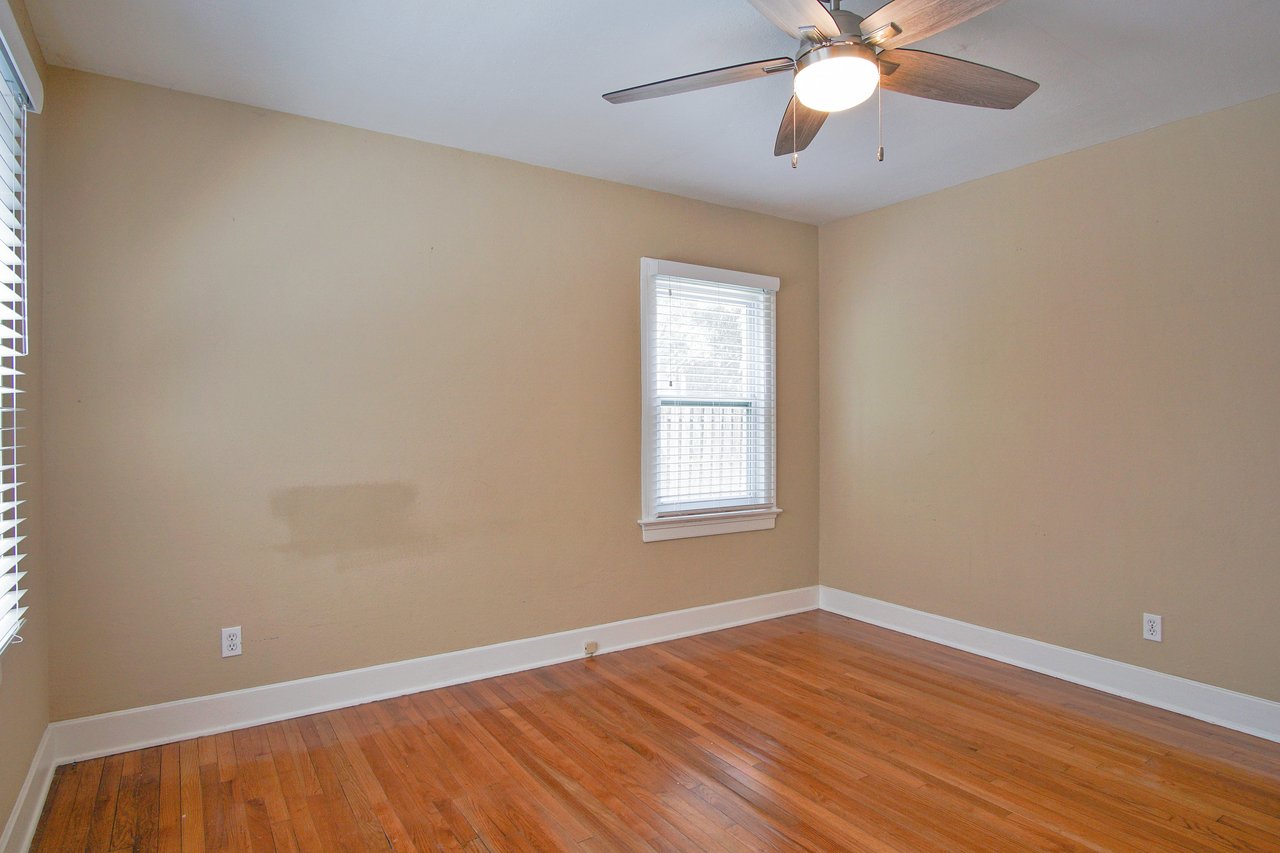 Empty room with beige walls, hardwood floor, large window with blinds, and a ceiling fan. The light creates a warm, inviting atmosphere.