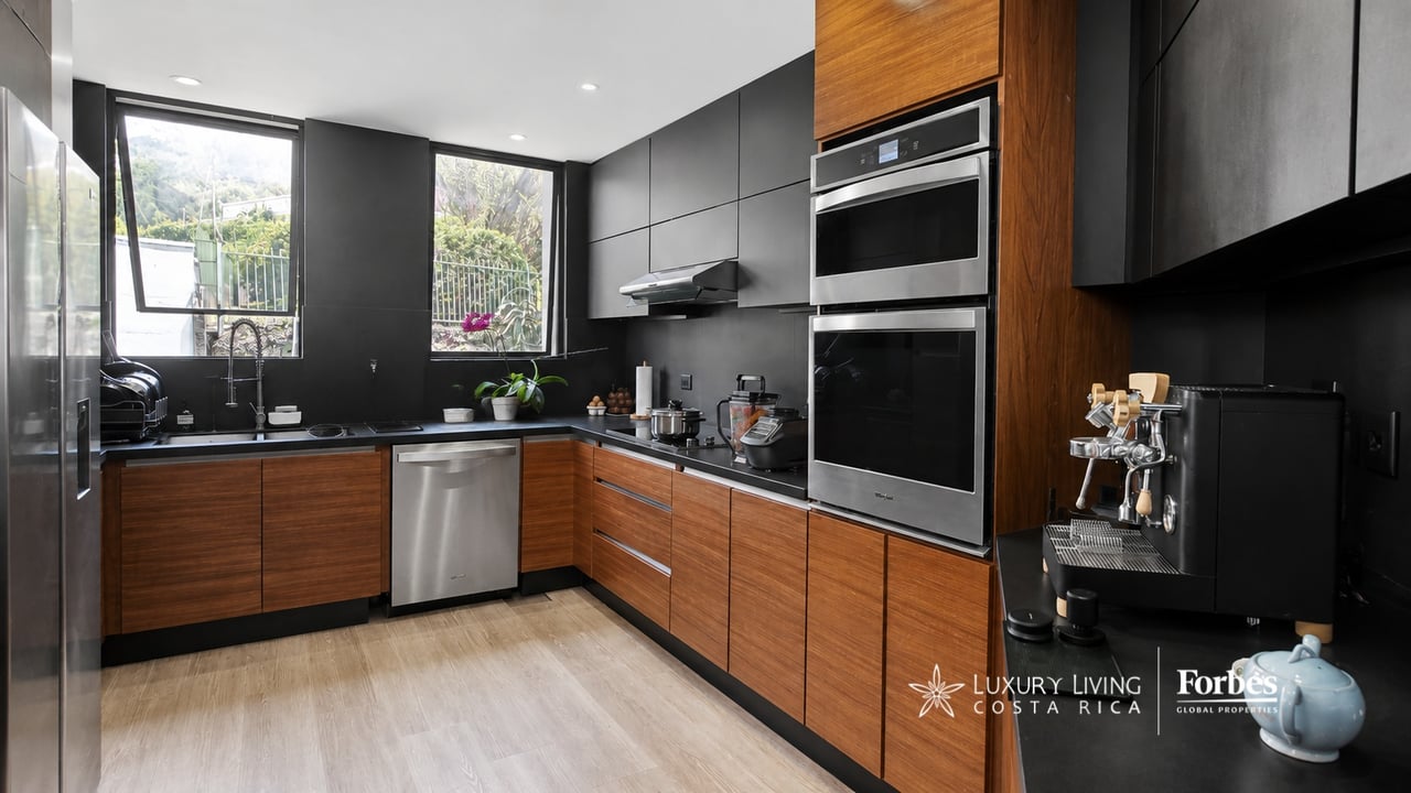 Modern kitchen with dark cabinetry, warm wood cabinets, stainless steel appliances, and windows overlooking greenery.